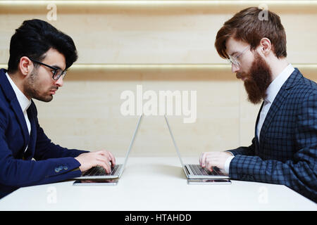 Profile view of two concentrated bearded businessmen in eyeglasses and suits sitting opposite one another and working on their laptops in office lobby Stock Photo