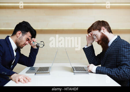 Two tired businessmen in stylish suits sitting at desk opposite one another and working on laptops, black-haired man holding eyeglasses in hand and ru Stock Photo