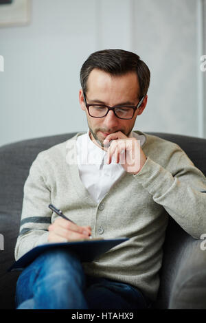 Pensive author or specialist sitting in armchair and making notes Stock Photo