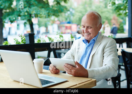 Portrait of stylish mature man using modern devices, laptop and tablet, at outdoor seating area in cafe, watching something and smiling happily on sum Stock Photo