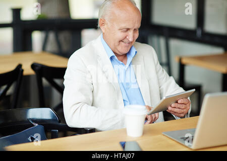 Modern senior man relaxing at outdoor area of city cafe while working with gadgets, laptop, smartphone and tablet, looking at screen and smiling Stock Photo