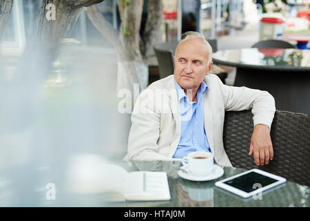 Portrait of quiet senior man sitting alone in street cafe, looking away pensively while having coffee break, with notebook and digital tablet laid out Stock Photo