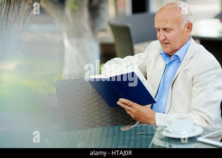 Portrait of elegant mature man looking into book while having coffee break at outdoor area of quiet cafe in shade of tree on summer day Stock Photo