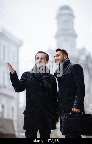 Image of stylish businessman showing the city to his international partner from middle east, standing in snowy street gesturing and smiling, discussin Stock Photo