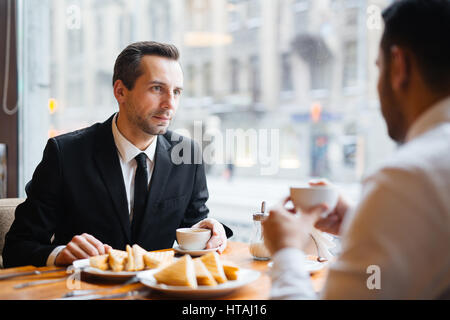 Portrait in warm lighting of two businessmen in formalwear discussing important matters in cafe over a light meal, focus on intent dark-haired man lis Stock Photo