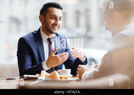Portrait of enthusiastic young Middle Eastern man gesturing actively and smiling talking to his business partner or colleague in quiet warm lit cafe o Stock Photo