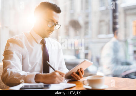 Young Middle Eastern business executive working at window table in cafe by rays of warm sunset light: holding tablet while making notes with pen on pa Stock Photo