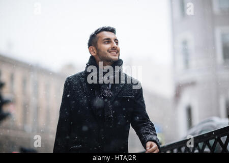 Waist up portrait of confident stylish Middle Eastern man in winter city: his coat covered with light snow as he is looking around smiling brightly Stock Photo