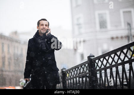 Portrait of stylish man, wearing coat and holding briefcase, talking on phone smiling happily while walking in snowy streets of big city Stock Photo