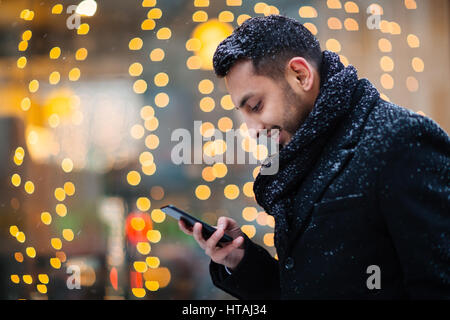 Heartwarming image of young Arabic man stopping to read pleasant text message in the streets of winter city on background of store window decorated by Stock Photo