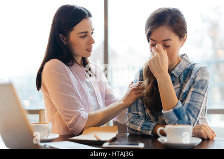 Sad young woman hiding her face crying at table in cafe and her friend hugging her by shoulders in compassion, giving advice trying to console her Stock Photo