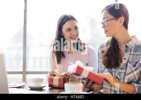 Sincere scene of two young women, one Asian, sharing gifts and talking at table in cafe sitting against big window, both smiling and looking caringly  Stock Photo