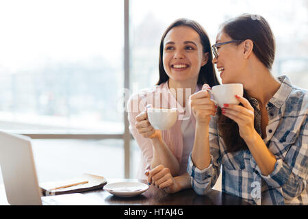 Portrait of two pretty young women, one of them Asian, laughing and talking during coffee break at work in office, holding big cups in hands and smili Stock Photo