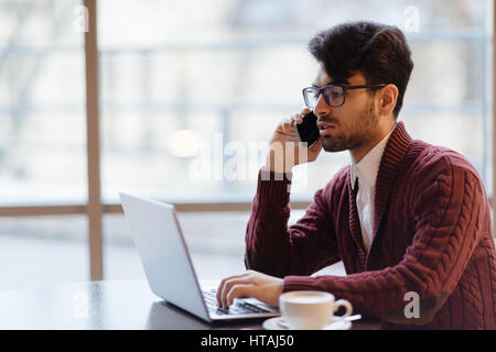 Profile view portrait of young Arabic businessman sitting at table against big window consulting with his partner about business deal via phone and ch Stock Photo