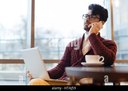 Modern middle-eastern businessman wearing knit sweater and black rim glasses busy working during coffee break in cafe against window, he is talking on Stock Photo