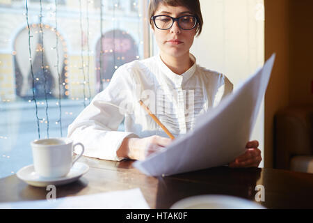 Portrait of serious elegant businesswoman wearing white shirt and glasses looking through paperwork while sitting in cafe at window table in evening Stock Photo