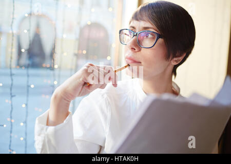 Head and shoulders portrait of young businesswoman looking away to window in thinking while sitting in cafe working with business papers Stock Photo
