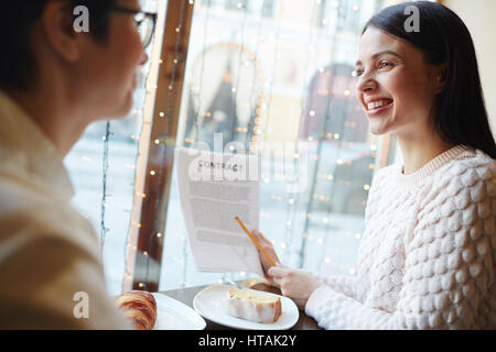 Portrait of young smiling woman presenting contract to interested business partner  during negotiations meeting  at cafe table in evening Stock Photo