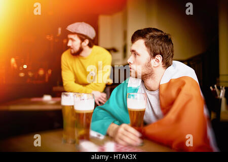 Side view of two men, one of them wrapped in Irish flag, looking away watching something on TV in Sports Pub with tall glasses of beer Stock Photo