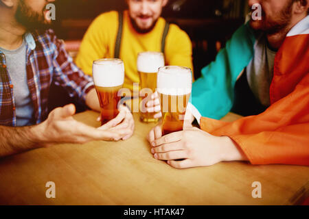 Closeup shot of tall glasses of lager beer in hands of three men sitting round table in Irish pub enjoying conversation Stock Photo