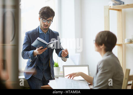 Portrait of contemporary young manager standing against window explaining work tasks to female colleague reading notes from planner book Stock Photo