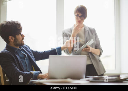 Young successful middle eastern man looking up from laptop to  consult young colleague standing by him with tablet, pointing at it Stock Photo