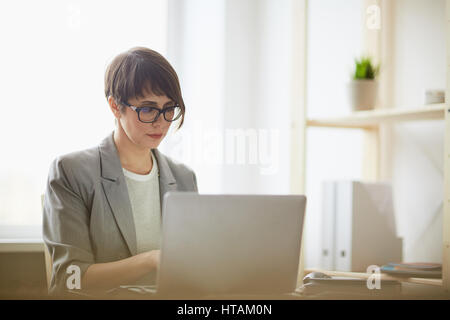 Portrait of young successful businesswoman wearing creative haircut and glasses busy typing something while working with laptop at workplace in modern Stock Photo