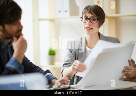 Portrait of business expert wearing creative haircut and glasses showing paperwork and explaining ideas to partner listening intently in modern office Stock Photo