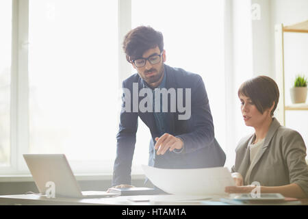 Successful young businesswoman showing plans to colleague leaning at her desk and discussing project Stock Photo