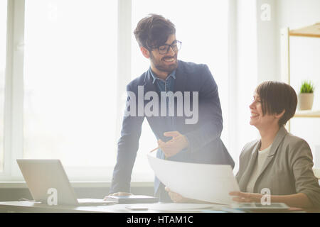 Portrait of  male manager praising young colleague leaning at her desk, pointing at working plans and smiling Stock Photo