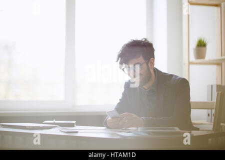 Sunlit portrait of contemporary middle eastern business man sitting at workplace in office and looking at smartphoe laughing  typing messages during b Stock Photo