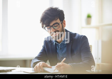 Sunlit portrait of young creative mn in glasses and business casual wear writing something down to notebook, smiling and looking pleased sitting at de Stock Photo