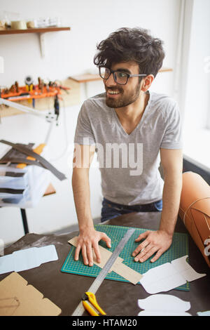 Portrait of muscular middle eastern man wearing creative haircut and glasses looking away and smiling, leaning on workshop table while creating leathe Stock Photo