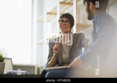 Two cheerful colleagues smiling while discussing something in modern office, focus on stylish businesswoman wearing creative haircut and glasses Stock Photo