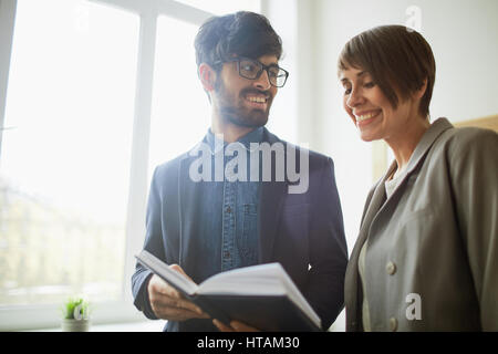 Two happy young business people, man and woman, wearing formalwear smiling and looking at opened planner book in sunlit modern office Stock Photo