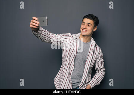 Cheerful selfie. Cheerful young man in shirt holding mobile phone and making photo of himself while standing against grey background Stock Photo