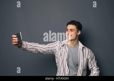 Cheerful selfie. Cheerful young man in shirt holding mobile phone and making photo of himself while standing against grey background Stock Photo
