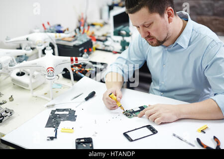 Portrait of modern man in workshop busy working on different electronic devices phones and drones on table with different tools and parts Stock Photo