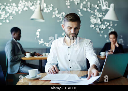 Portrait of young bearded business man reading documents while working with laptop in cafe during coffee break, two people behind him Stock Photo