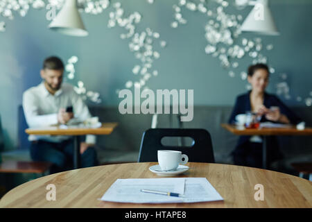 Image of business documents on empty table in cafe with two businesspeople working in background during coffee break Stock Photo