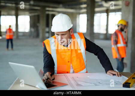 Portrait of development supervisor wearing protective vest and helmet over formal suit  proofing blueprints using laptop computer inside unfinished bu Stock Photo