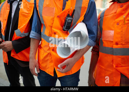 Closeup shot of three unrecognizable construction workers wearing orange reflective vests standing on site holding building plans Stock Photo