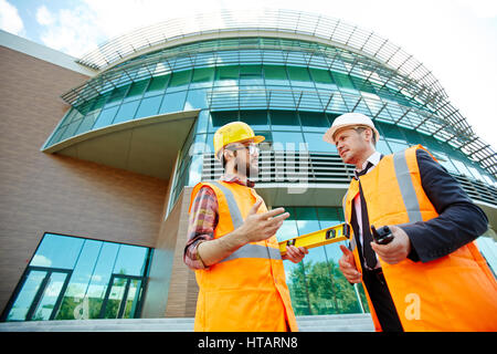 Low angle view of two business people wearing protective vests and hard hats standing on construction site, executive supervisor talking to young arch Stock Photo