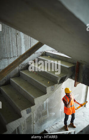 High angle view of construction worker wearing reflective orange vest and protective helmet standing using portable radio under unfinished flight of s Stock Photo
