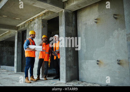 Two workmen showing unfinished floors in high rise building to executive supervisor on construction site, all wearing reflective orange vests and hard Stock Photo