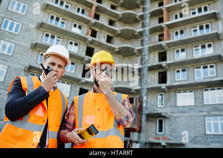 Portrait of two workmen wearing reflective orange vests and hard hats standing using portable radio sets against apartment building on construction si Stock Photo