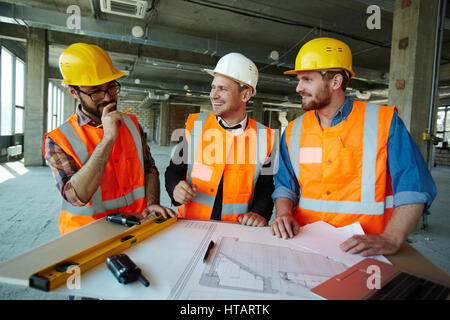 Team of cheerful construction workers wearing protective helmets and vests smiling and laughing while discussing project details with executive superv Stock Photo