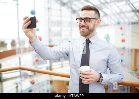 Portrait of contemporary bearded businessman wearing shirt and glasses video chatting using smartphone during coffee break on stairs of office buildin Stock Photo