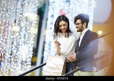 Portrait of elegant couple in business center: young stylish man and woman going down the escalator with bags, girlfriend showing pictures in smartpho Stock Photo