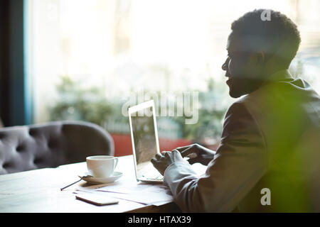 Side view portrait of successful African- American businessman wearing formal suit looking at screen while busy working with laptop in cafe Stock Photo